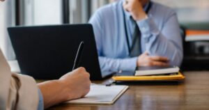 A job applicant filling in a paper application form across a desk from an interviewer, beside a laptop