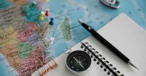 Paper world map with coloured pins, a compass, a blank spiral notebook and a pencil laid out for trip planning.