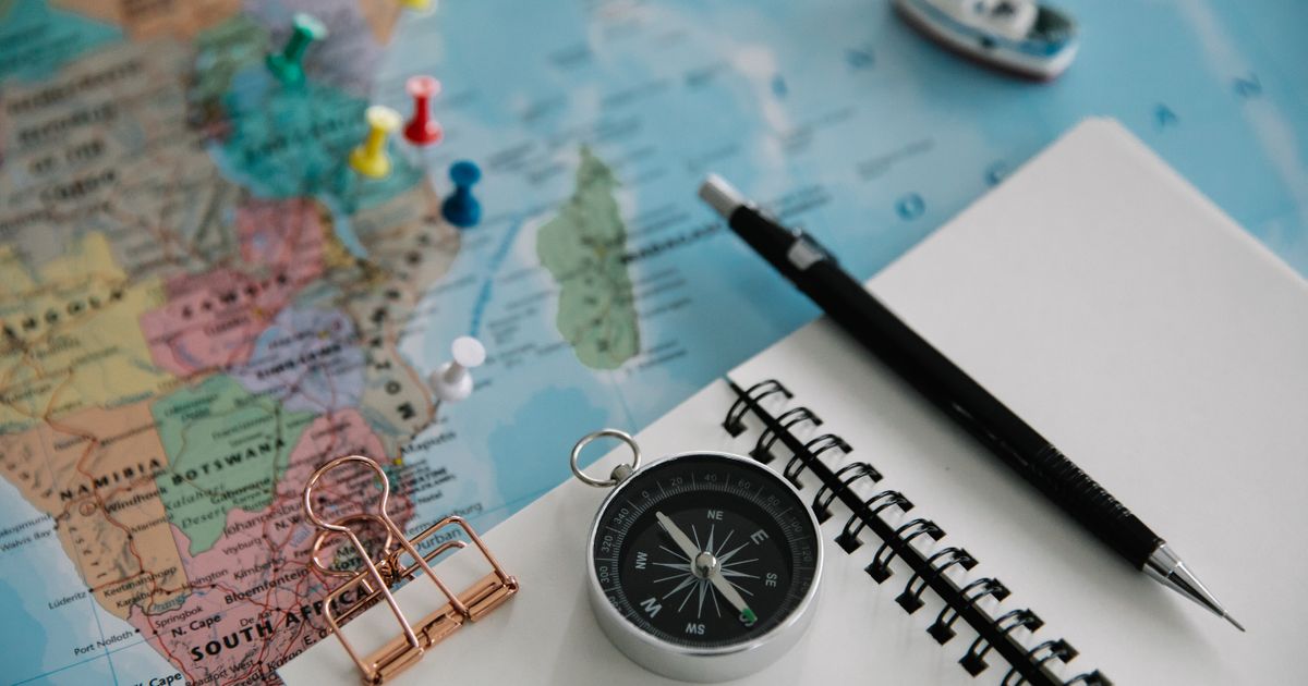 Paper world map with coloured pins, a compass, a blank spiral notebook and a pencil laid out for trip planning.