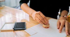 Hands pressing a wooden rubber stamp onto a sheet of paper beside an ink pad, illustrating how certification and credential claims must be verified before they are advertised in Singapore.