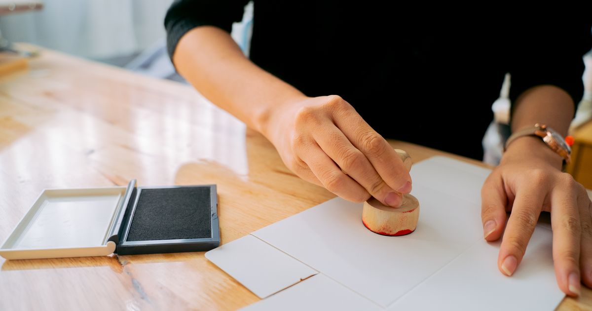 Hands pressing a wooden rubber stamp onto a sheet of paper beside an ink pad, illustrating how certification and credential claims must be verified before they are advertised in Singapore.