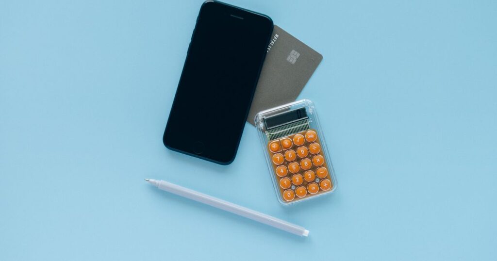 A smartphone with a blank screen, an unbranded payment card and an orange calculator on a pale blue desk, illustrating credit card, loan and BNPL advertising rules in Singapore.