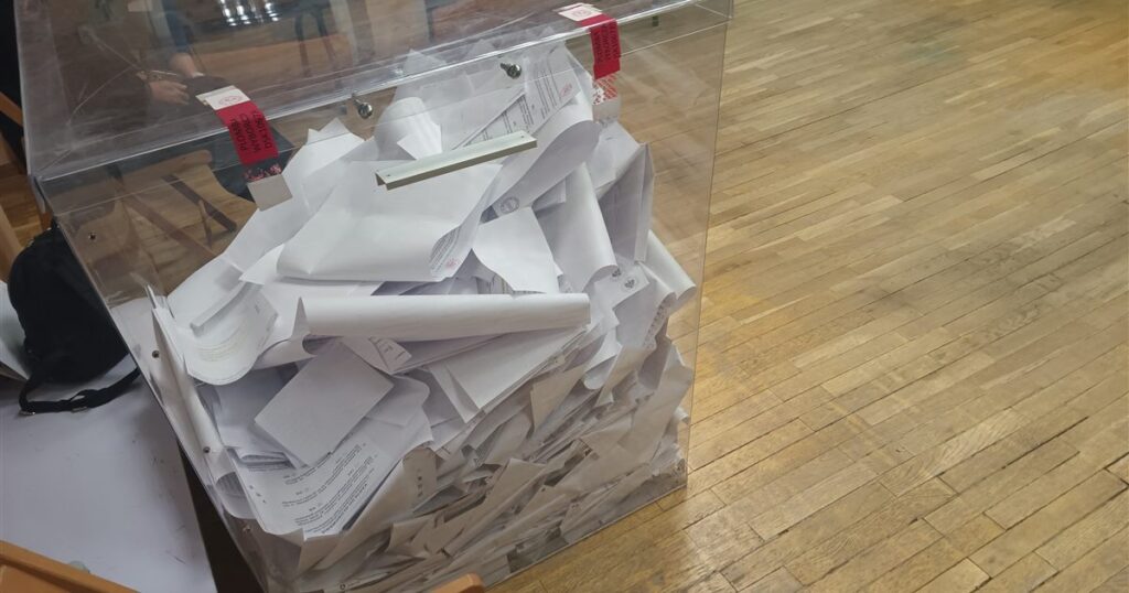 A clear acrylic ballot box filled with folded paper ballots on a wooden floor, illustrating the rules on election advertising for brands and agencies in Singapore.