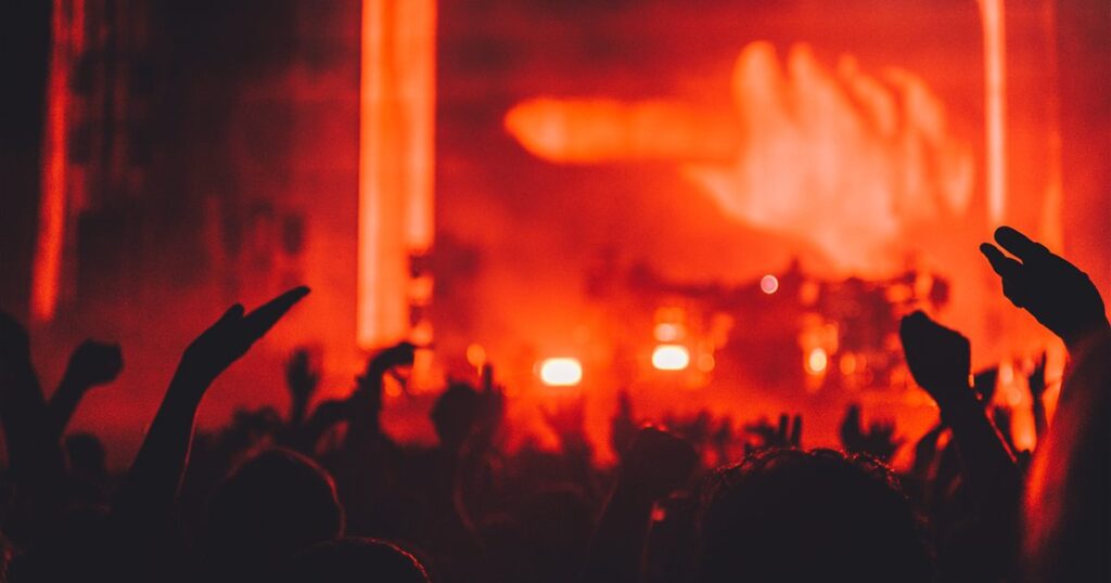 Silhouetted concert crowd with raised hands in front of a stage washed in red light, illustrating the licensing and publicity rules for marketing public events in Singapore.
