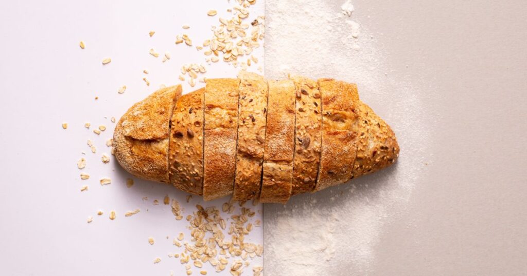 A sliced wholegrain loaf with scattered oats and flour on a white and grey background, illustrating the rules on using the Healthier Choice Symbol in Singapore food marketing.