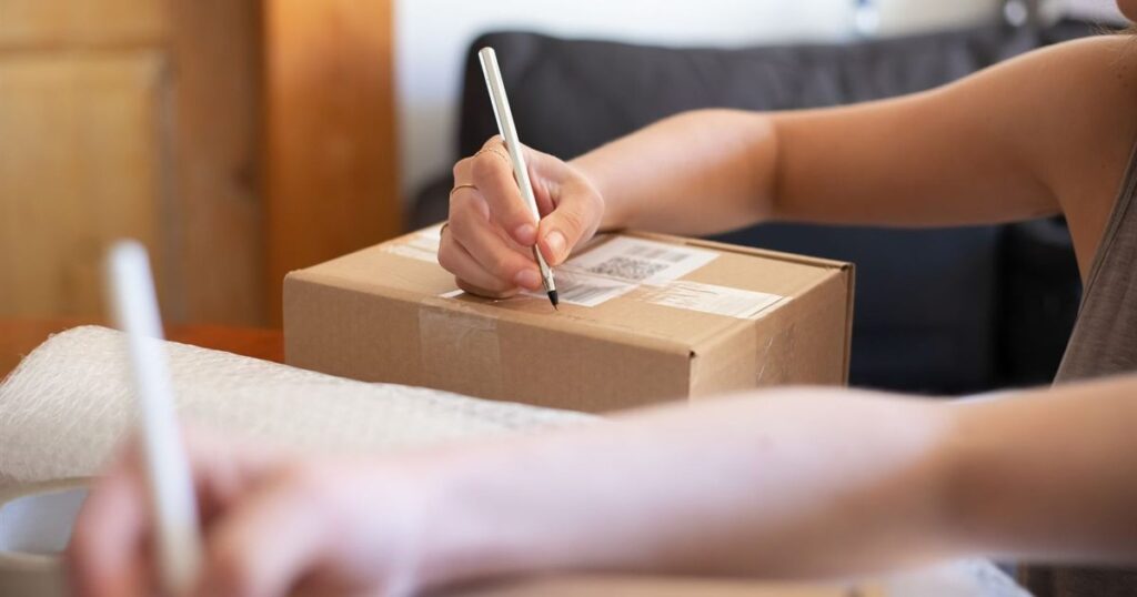 Hands writing on a labelled cardboard parcel at home next to bubble wrap, illustrating the rules for marketing and selling from a home-based business in Singapore.