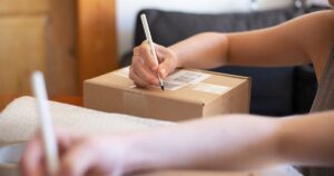 Hands writing on a labelled cardboard parcel at home next to bubble wrap, illustrating the rules for marketing and selling from a home-based business in Singapore.