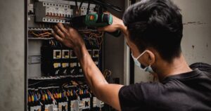 An electrician in a dark T-shirt using a cordless drill on wiring inside an open electrical distribution panel, illustrating licensing rules for advertising electrical work in Singapore.