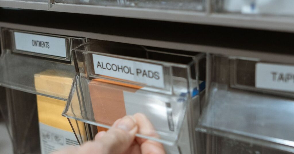 A hand opening a labelled clear drawer of alcohol pads beside ointments in a pharmacy supply cabinet, illustrating the rules on advertising by pharmacies in Singapore.