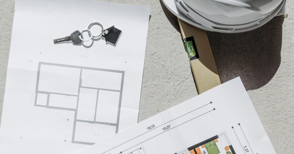 Printed floor plans, a house-shaped keyring, a spirit level and a white hard hat on concrete, illustrating advertising rules for renovation contractors in Singapore.